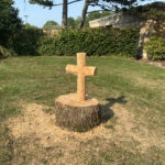An outdoor shot shows a tree stump in a grassy yard, which has been carved into a Christian cross. Sawdust is scattered on the grass around the base of the stump. In the background are two tall evergreen trees, a green hedge, and a light-colored building under a clear blue sky.